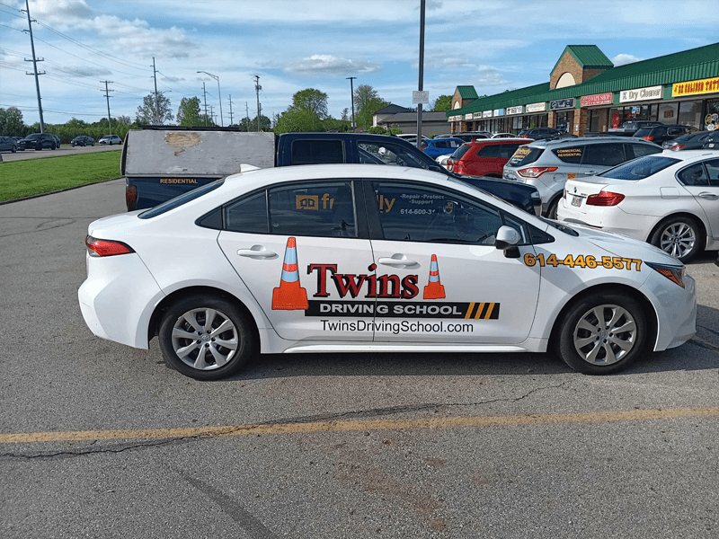 White car marked as "Twins Driving School" parked in a shopping plaza lot.